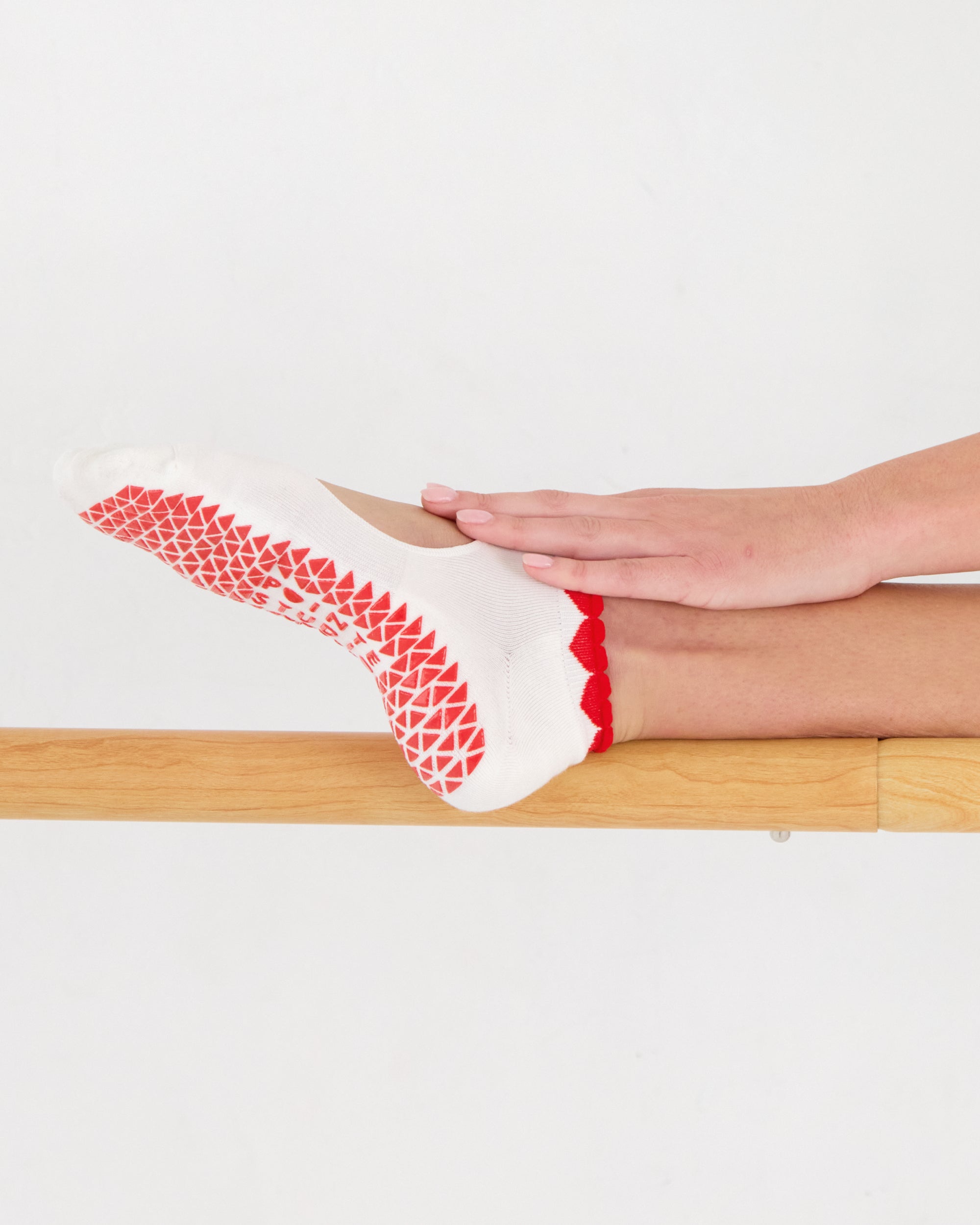 Woman on a barre wearing solid white strap grip sock with red heart wrapping around the scalloped cuff, made for pilates, barre, yoga, and beyond. 