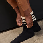 Women wearing a black mid-weight knit grip sock that scrunches with beige stripes on the ankle part, made for Pilates, Barre, Yoga, and Beyond. 