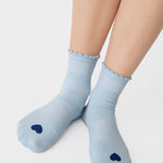 Women wearing a light blue ankle grip sock with a navy blue heart motif on the top made for Pilates, Barre, Yoga, and beyond. 