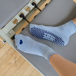 Women on a pilates reformer wearing a light blue ankle grip sock with a navy blue heart motif on the top made for Pilates, Barre, Yoga, and beyond. 