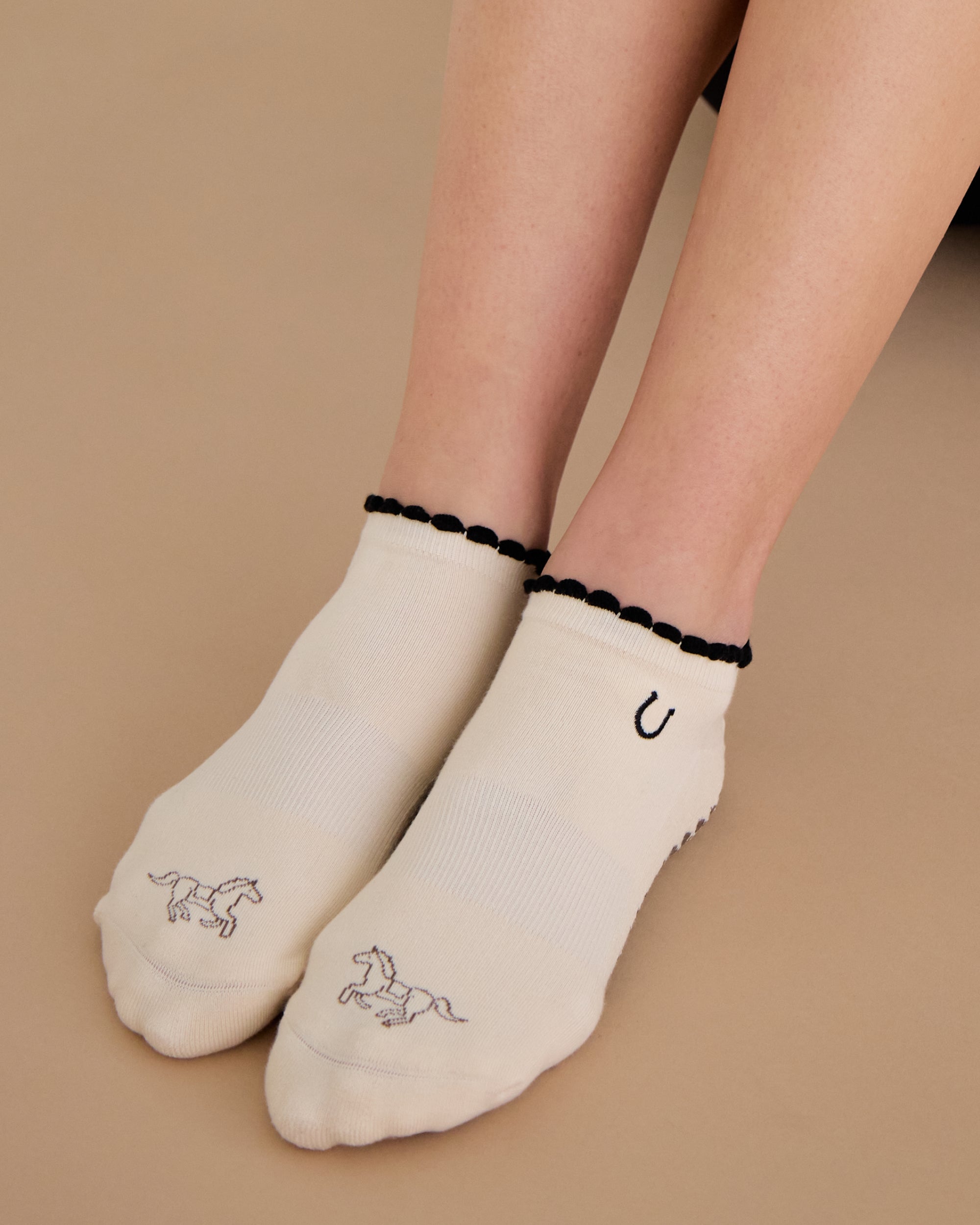 Woman wearing a beige full foot grip sock with a horse on the top and a horseshoe embroidered along the ankle. Made for pilates, barre, and yoga. 