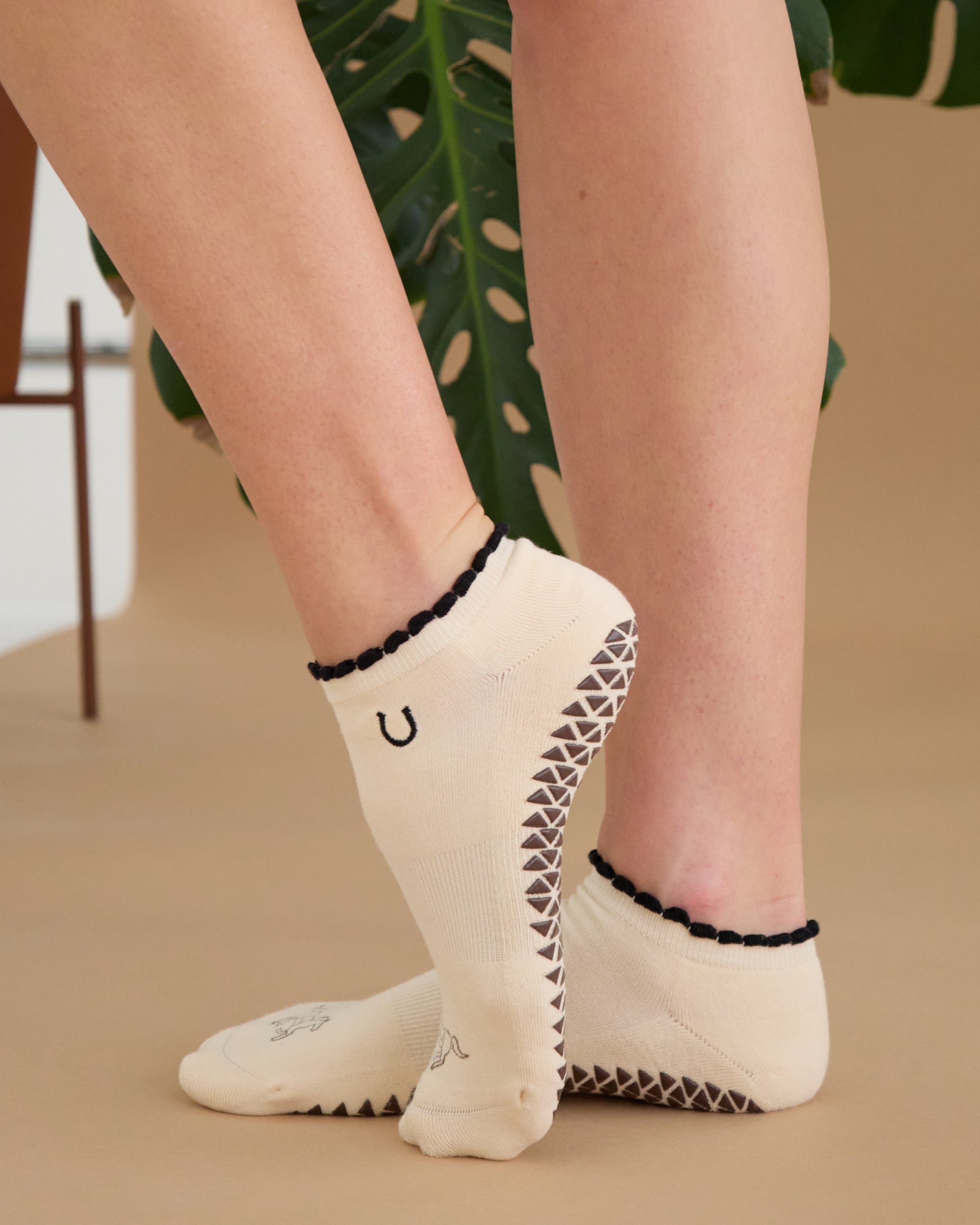 Woman wearing a beige full foot grip sock with a horse on the top and a horseshoe embroidered along the ankle. Made for pilates, barre, and yoga. 