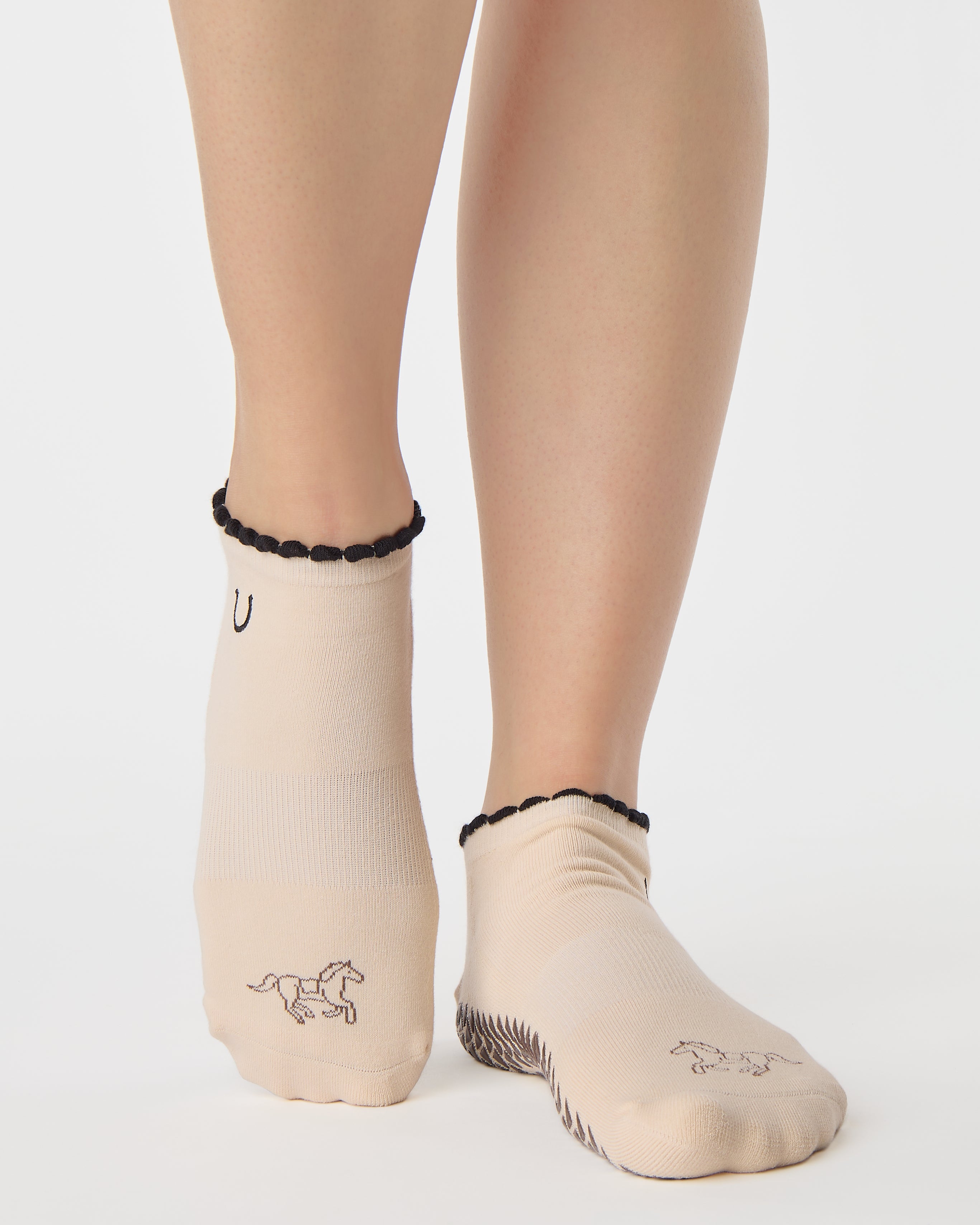 Woman wearing a beige full foot grip sock with a horse on the top and a horseshoe embroidered along the ankle. Made for pilates, barre, and yoga. 