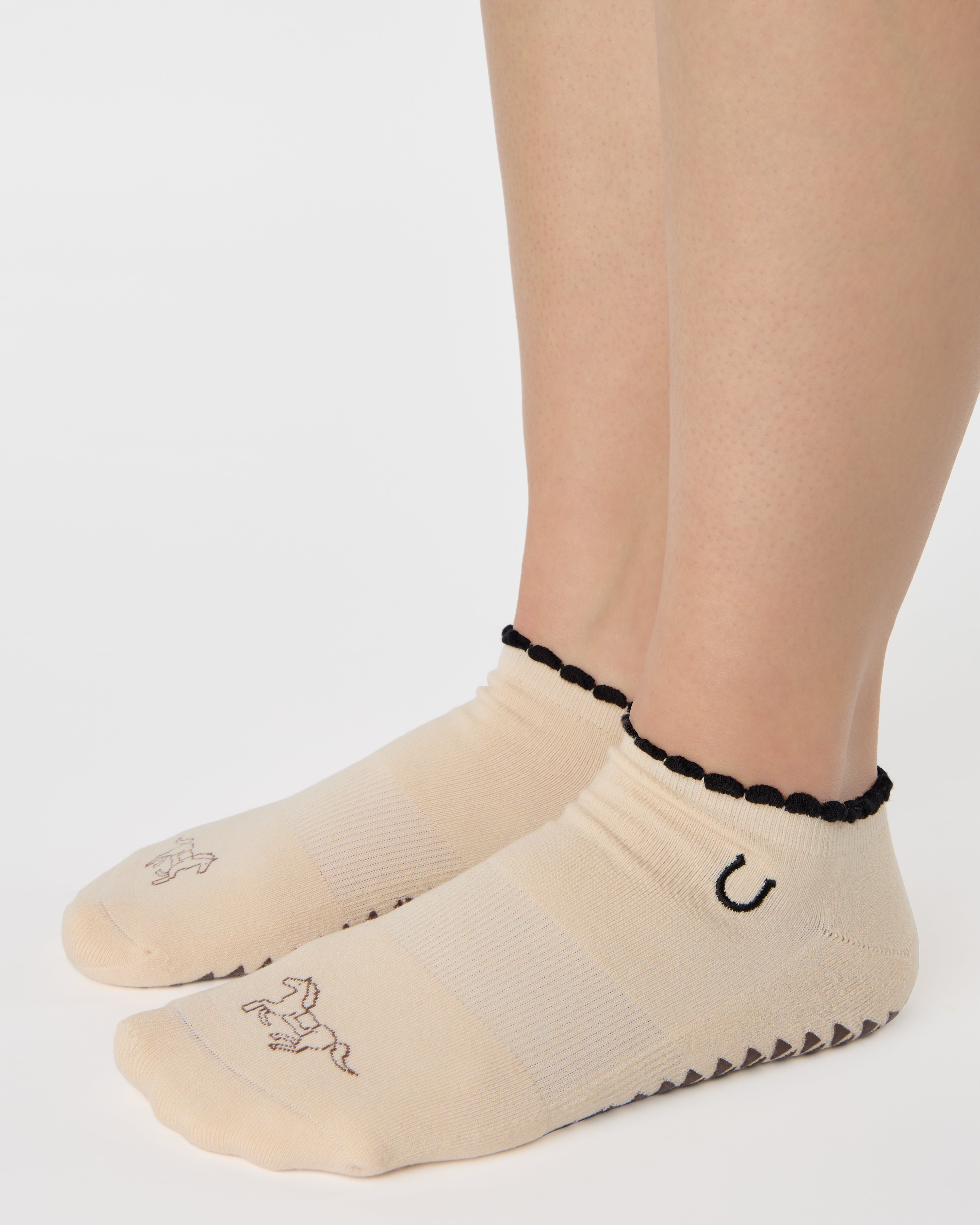 Woman wearing a beige full foot grip sock with a horse on the top and a horseshoe embroidered along the ankle. Made for pilates, barre, and yoga. 