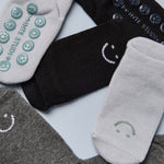 A grip sock pack of mini grip socks for kids ages 1-3, each pair has a happy face motif, black, charcoal and light blue styles.