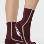 Woman wearing a brownish cherry red solid color ankle grip sock with two green and blue bold racing stripes on the side, made for pilates, barre, yoga, and beyond. 