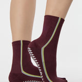 Woman wearing a brownish cherry red solid color ankle grip sock with two green and blue bold racing stripes on the side, made for pilates, barre, yoga, and beyond. 