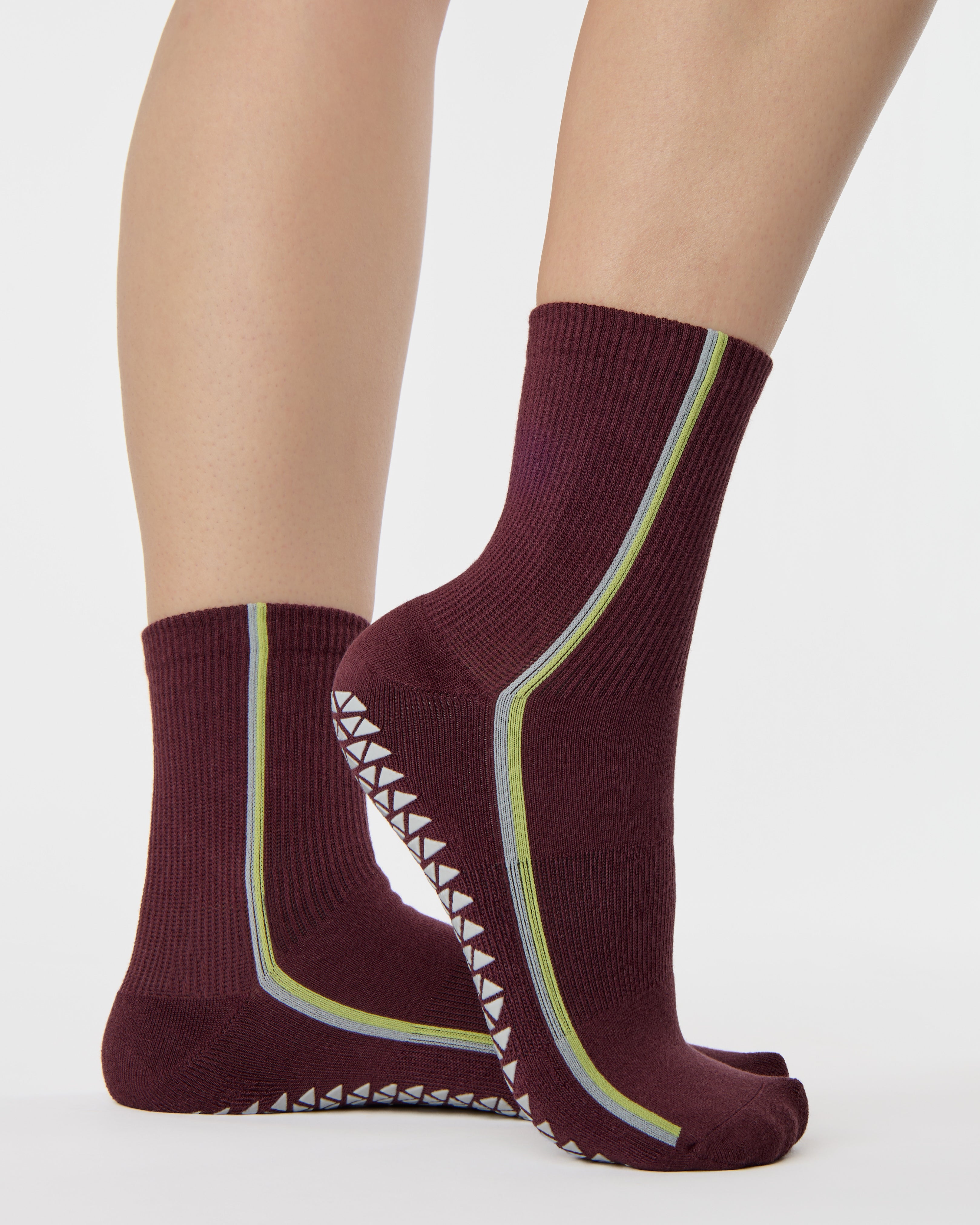 Woman wearing a brownish cherry red solid color ankle grip sock with two green and blue bold racing stripes on the side, made for pilates, barre, yoga, and beyond. 