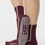 Woman wearing a brownish cherry red solid color ankle grip sock with two green and blue bold racing stripes on the side, made for pilates, barre, yoga, and beyond. 