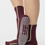 Woman wearing a brownish cherry red solid color ankle grip sock with two green and blue bold racing stripes on the side, made for pilates, barre, yoga, and beyond. 