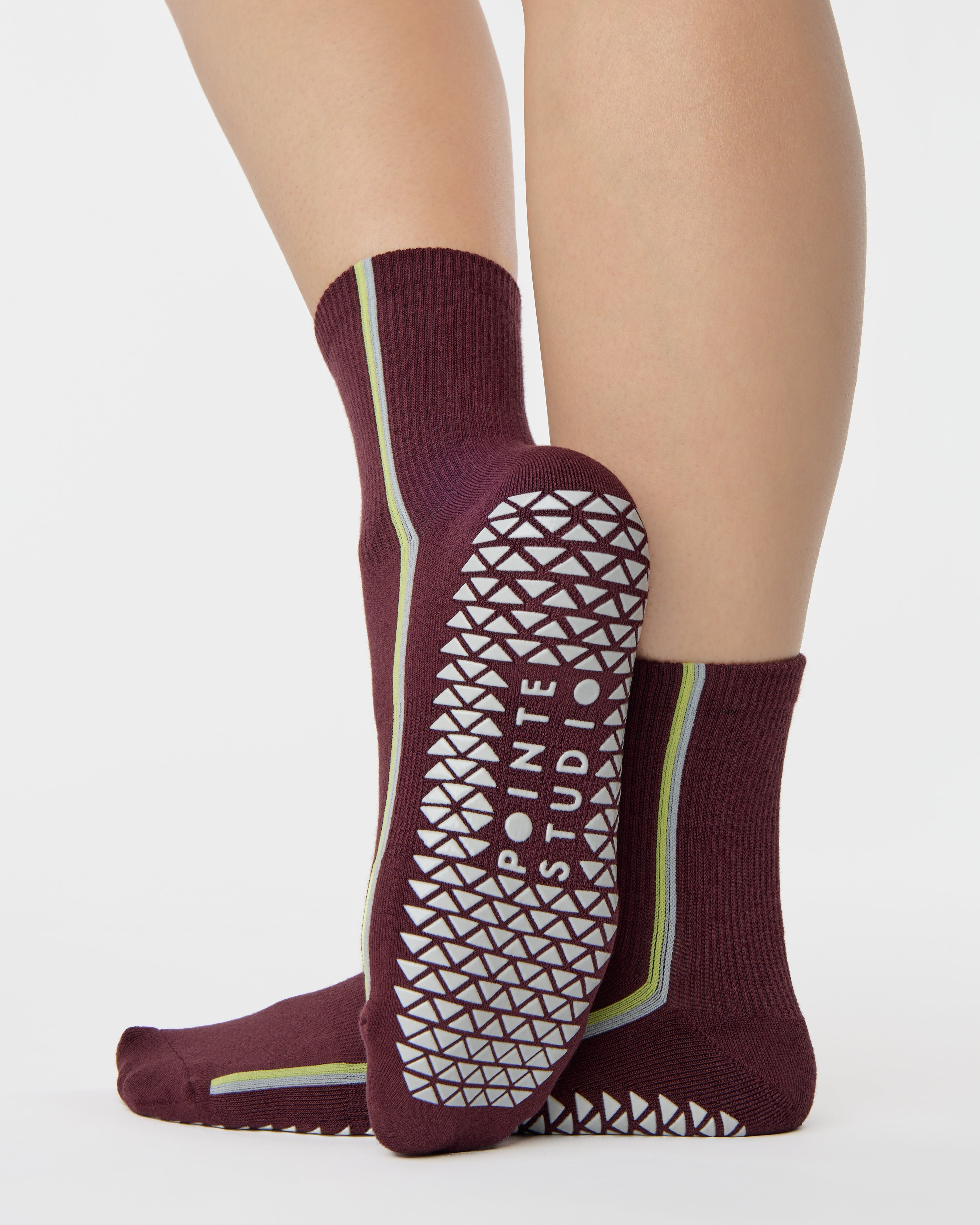 Woman wearing a brownish cherry red solid color ankle grip sock with two green and blue bold racing stripes on the side, made for pilates, barre, yoga, and beyond. 