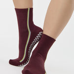 Woman wearing a brownish cherry red solid color ankle grip sock with two green and blue bold racing stripes on the side, made for pilates, barre, yoga, and beyond. 