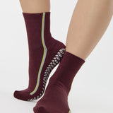 Woman wearing a brownish cherry red solid color ankle grip sock with two green and blue bold racing stripes on the side, made for pilates, barre, yoga, and beyond. 