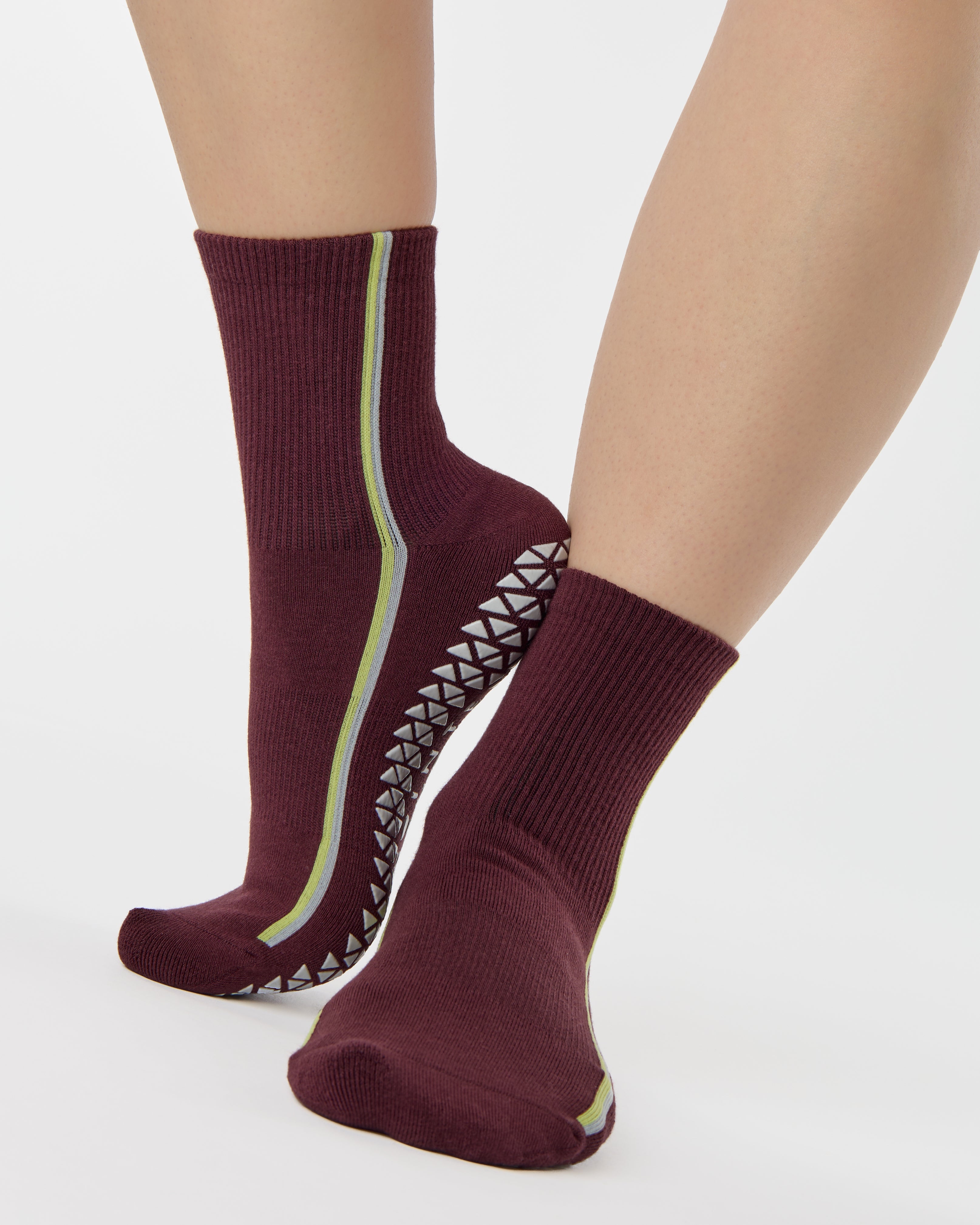 Woman wearing a brownish cherry red solid color ankle grip sock with two green and blue bold racing stripes on the side, made for pilates, barre, yoga, and beyond. 