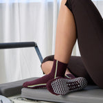 Woman on a pilates reformer wearing a brownish cherry red solid color ankle grip sock with two green and blue bold racing stripes on the side, made for pilates, barre, yoga, and beyond. 