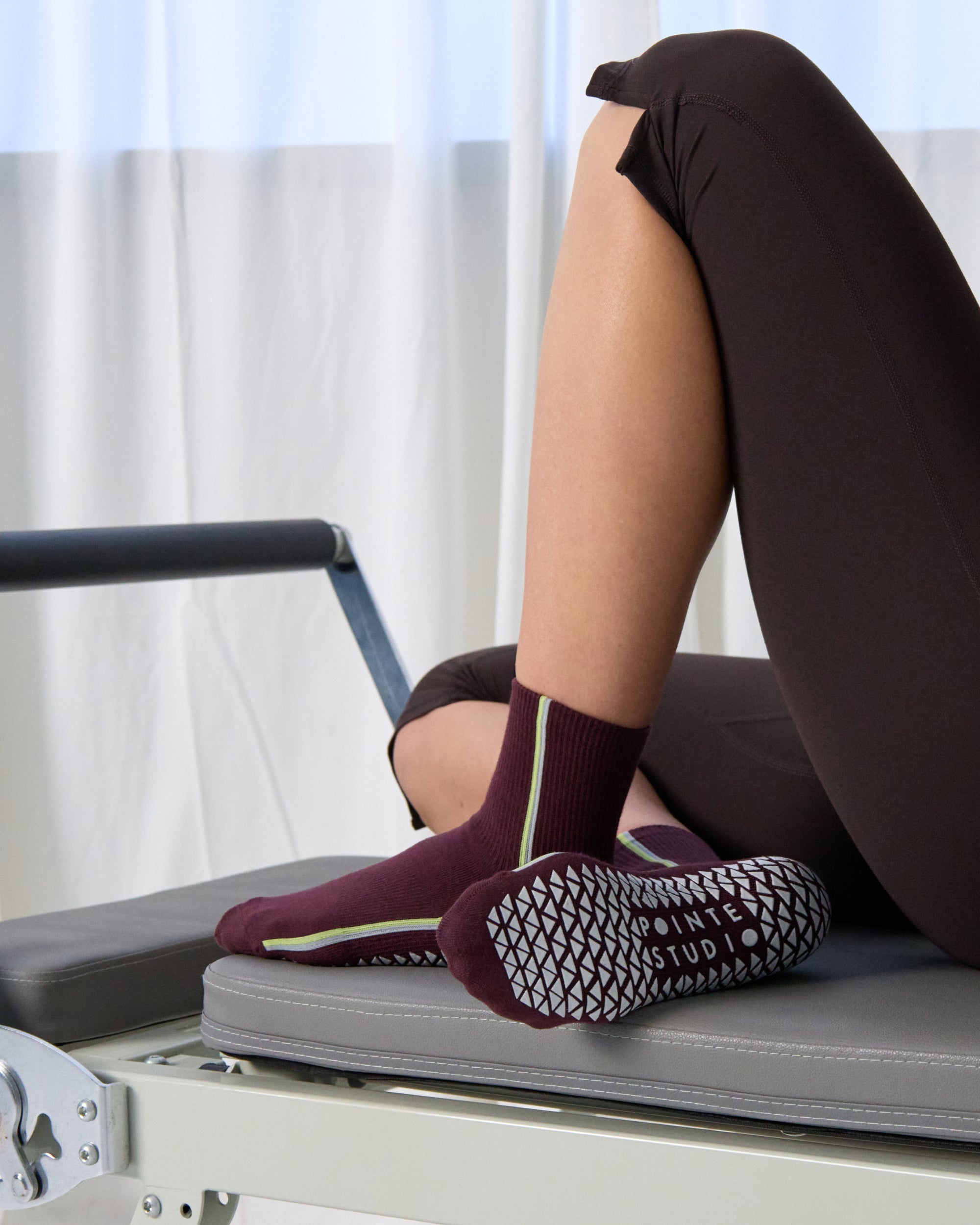 Woman on a pilates reformer wearing a brownish cherry red solid color ankle grip sock with two green and blue bold racing stripes on the side, made for pilates, barre, yoga, and beyond. 