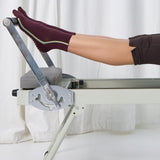Woman pilates reformer wearing a brownish cherry red solid color ankle grip sock with two green and blue bold racing stripes on the side, made for pilates, barre, yoga, and beyond. 