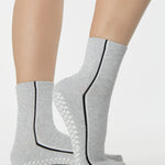 Woman wearing a heather grey solid color ankle grip sock with two black and white bold racing stripes on the side, made for pilates, barre, yoga, and beyond. 