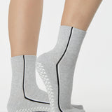 Woman wearing a heather grey solid color ankle grip sock with two black and white bold racing stripes on the side, made for pilates, barre, yoga, and beyond. 