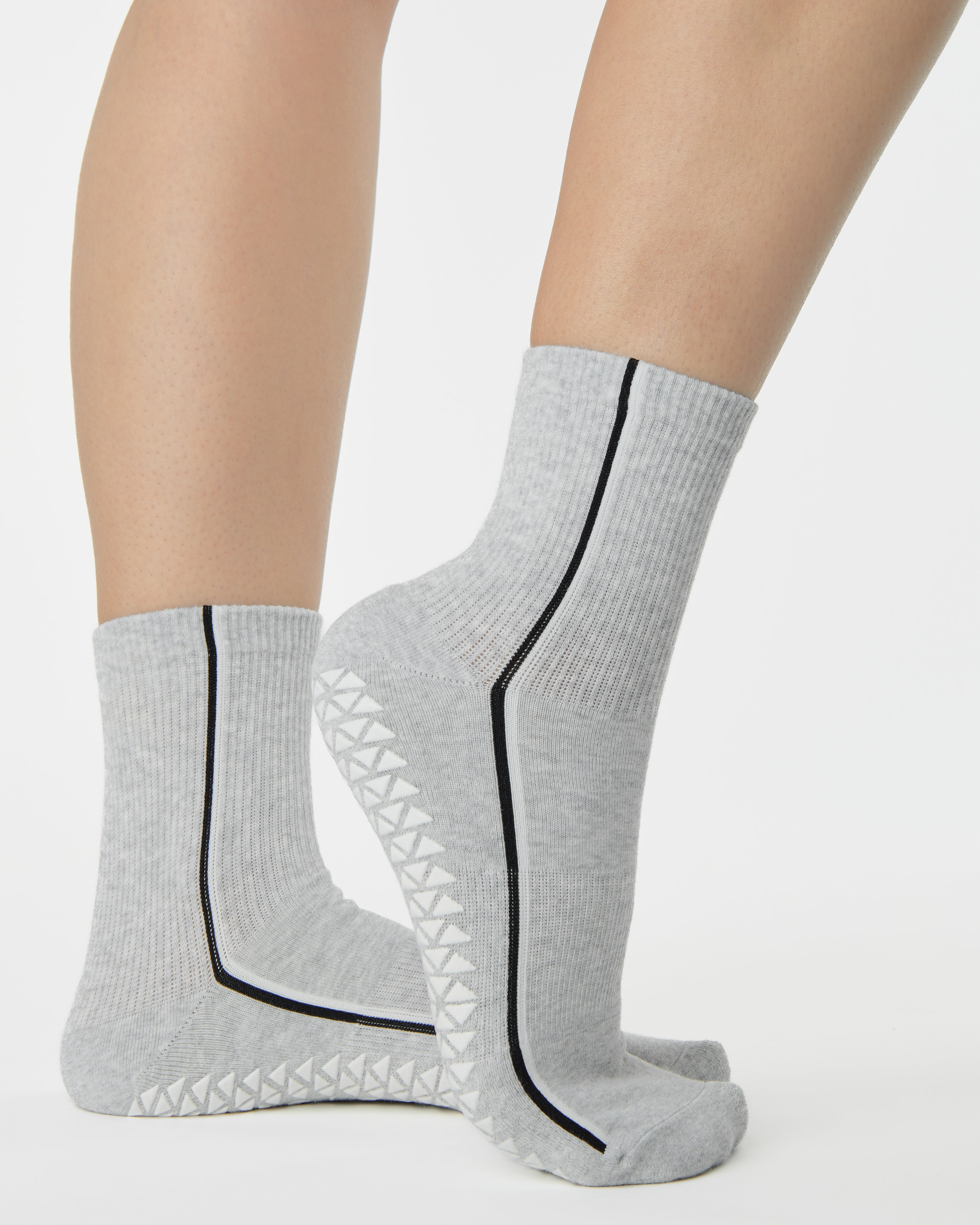 Woman wearing a heather grey solid color ankle grip sock with two black and white bold racing stripes on the side, made for pilates, barre, yoga, and beyond. 