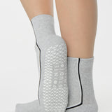 Woman wearing a heather grey solid color ankle grip sock with two black and white bold racing stripes on the side, made for pilates, barre, yoga, and beyond. 
