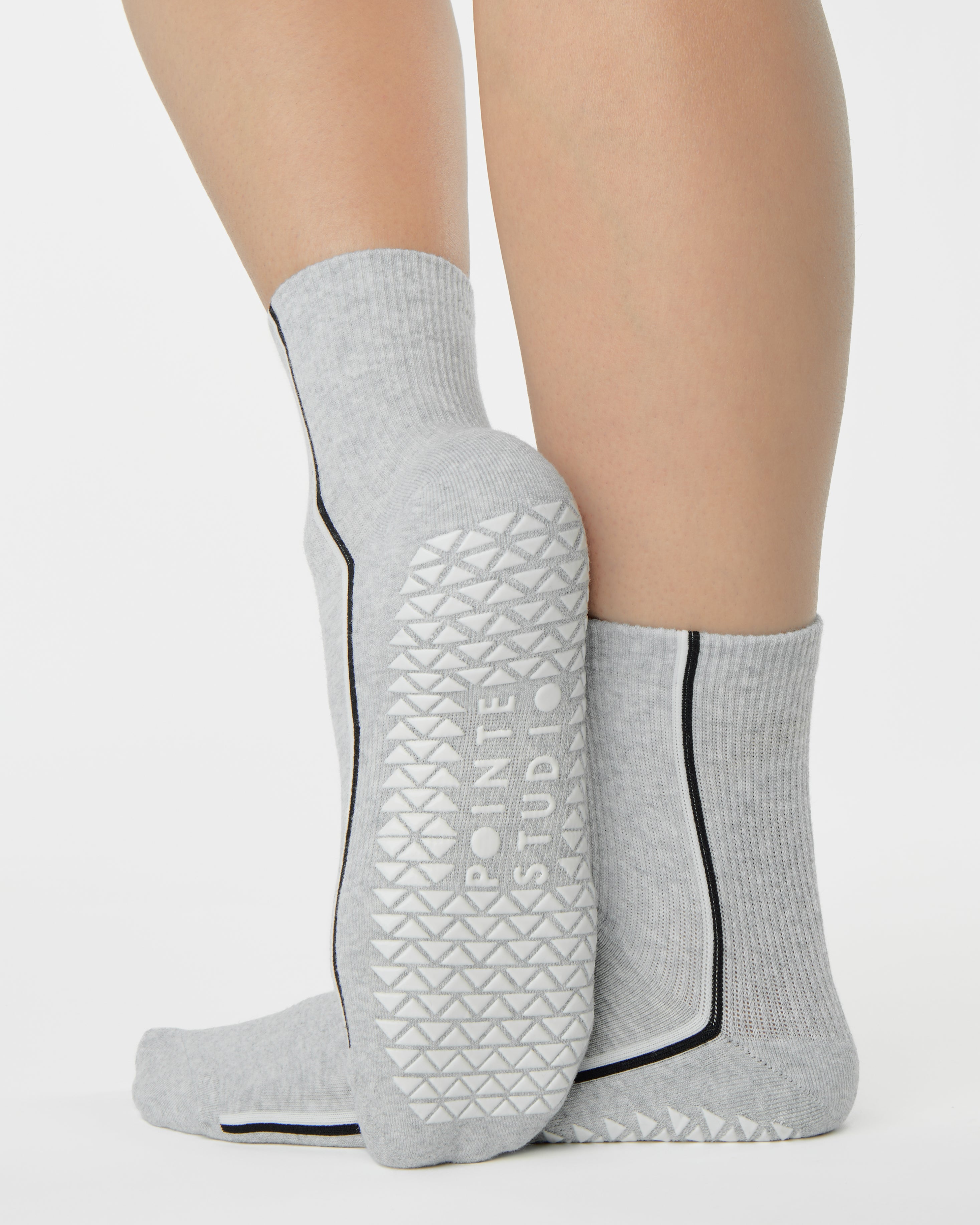 Woman wearing a heather grey solid color ankle grip sock with two black and white bold racing stripes on the side, made for pilates, barre, yoga, and beyond. 