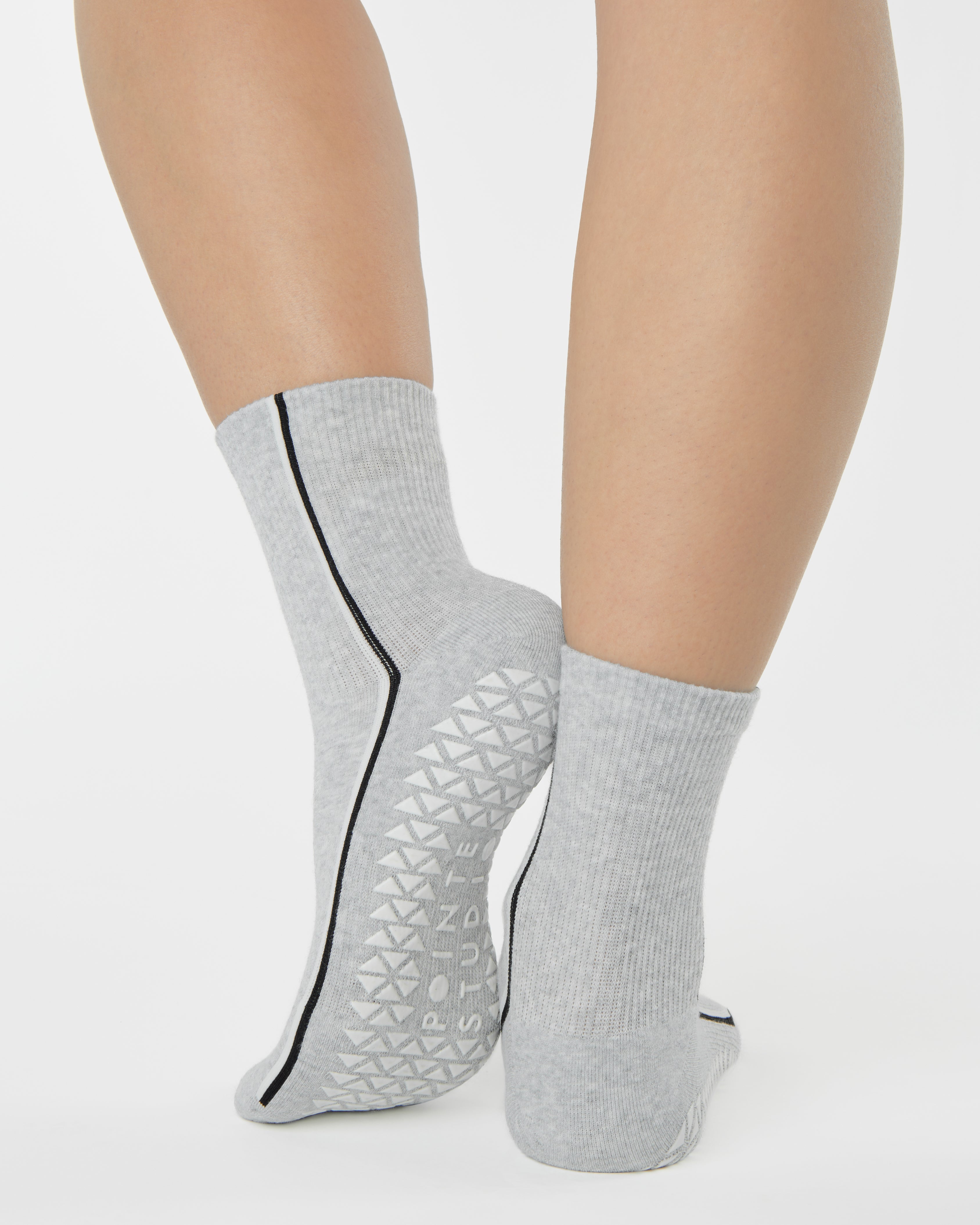 Woman wearing a heather grey solid color ankle grip sock with two black and white bold racing stripes on the side, made for pilates, barre, yoga, and beyond. 