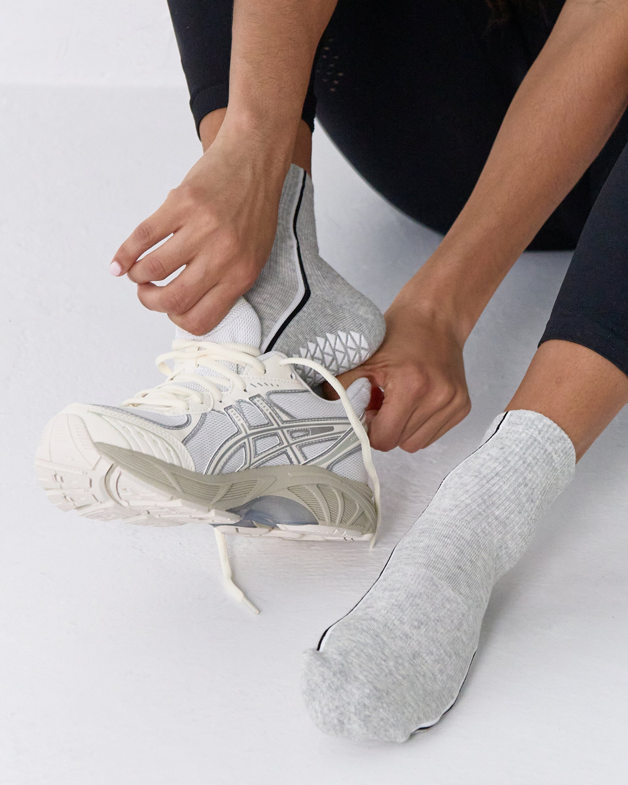 Woman wearing a heather grey solid color ankle grip sock with two black and white bold racing stripes on the side, made for pilates, barre, yoga, and beyond. 