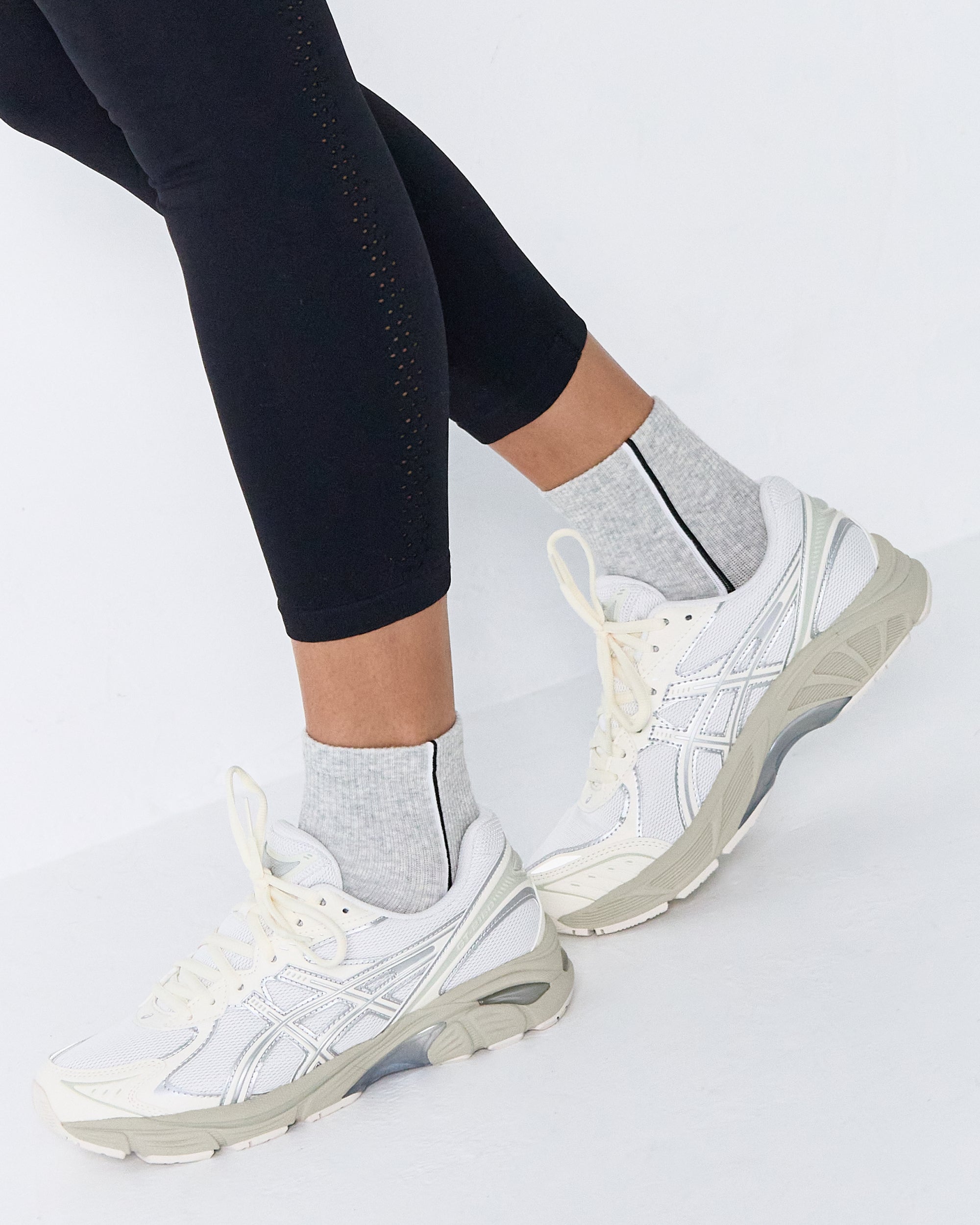 Woman wearing a heather grey solid color ankle grip sock with two black and white bold racing stripes on the side, made for pilates, barre, yoga, and beyond. 