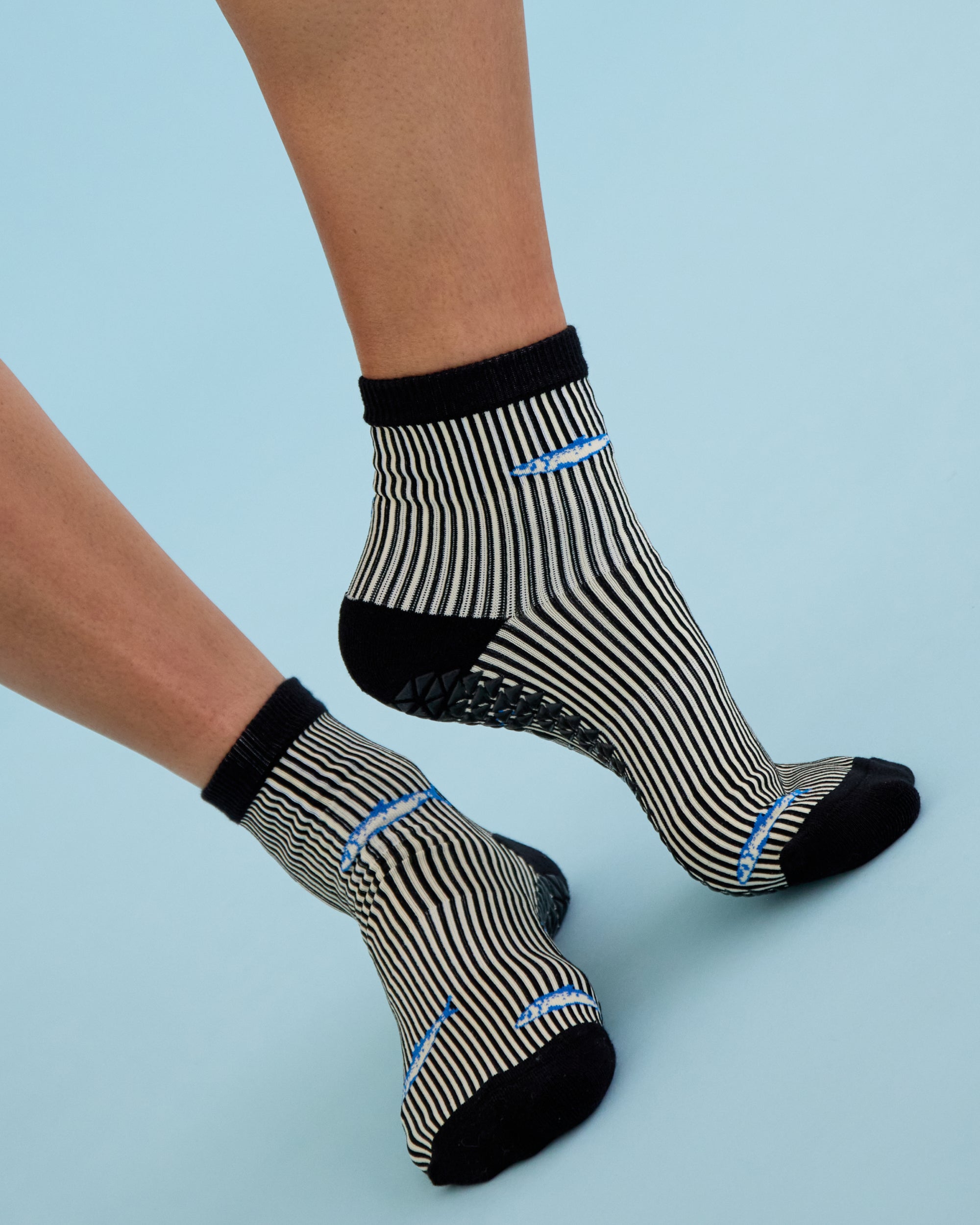 Woman wearing a black and white striped ankle grip sock with sardines swimming around the sock. Made for pilates, barre, yoga, and beyond. 