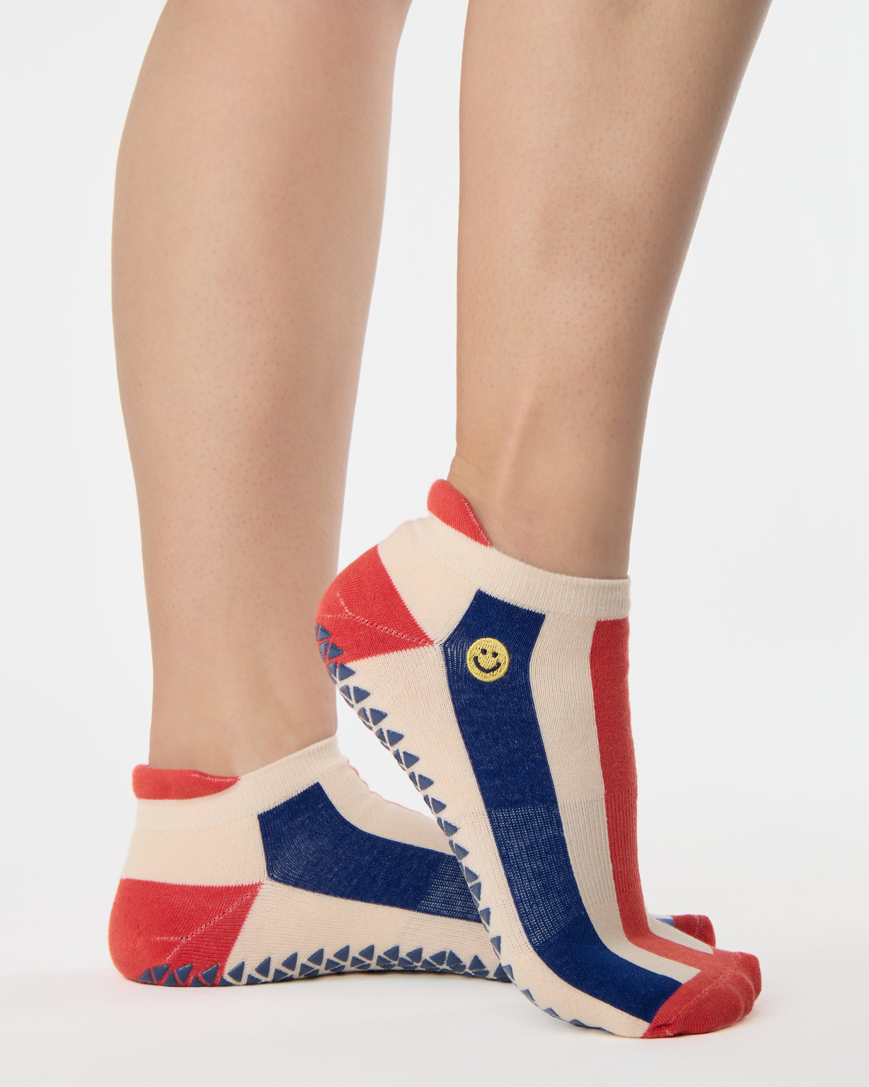 Woman wearing a playfully blue, white, and red stripped full foot grip sock with a little embroidered smiley face by the ankle. Made for pilates, barre, yoga, and beyond. 