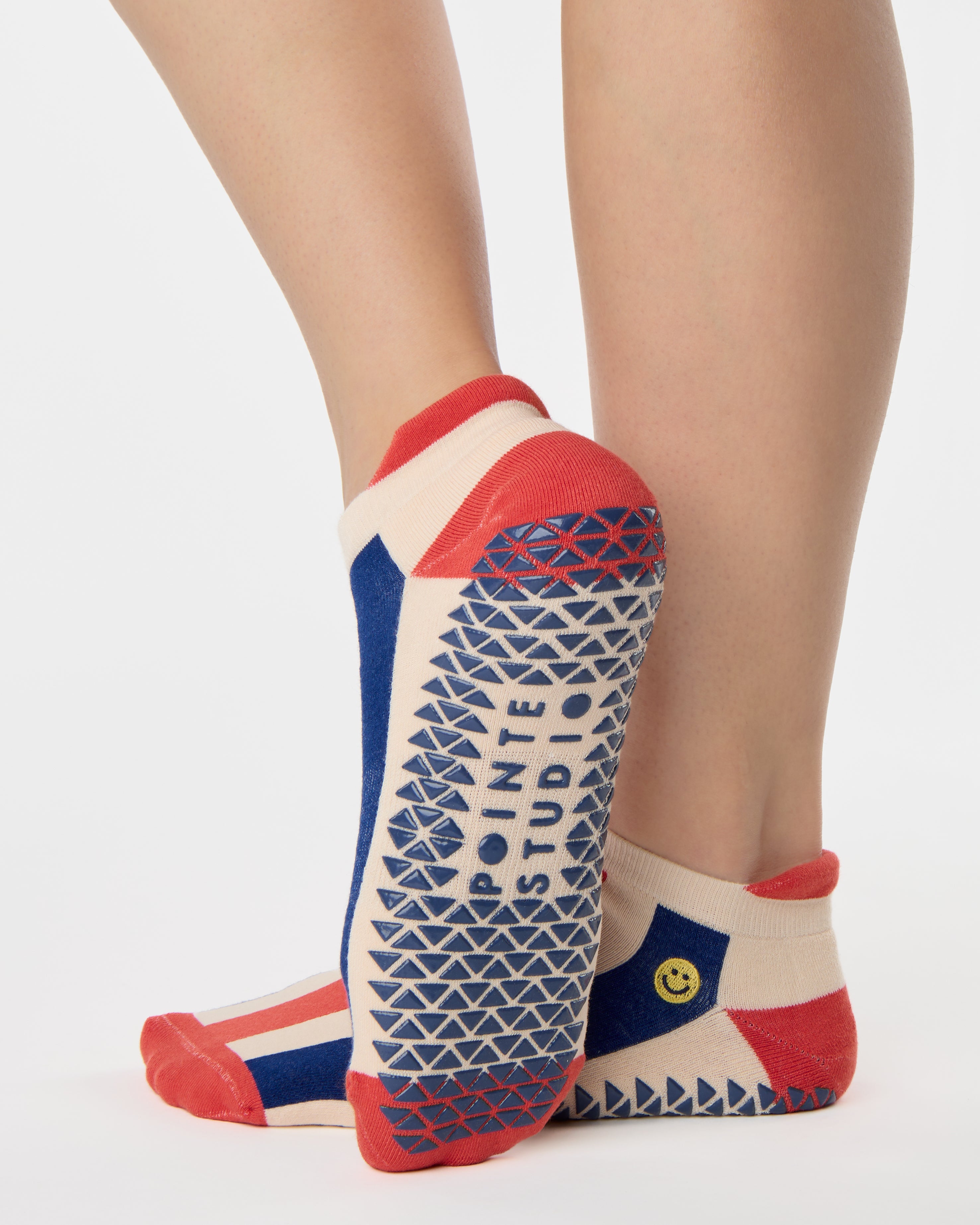 Woman wearing a playfully blue, white, and red stripped full foot grip sock with a little embroidered smiley face by the ankle. Made for pilates, barre, yoga, and beyond. 