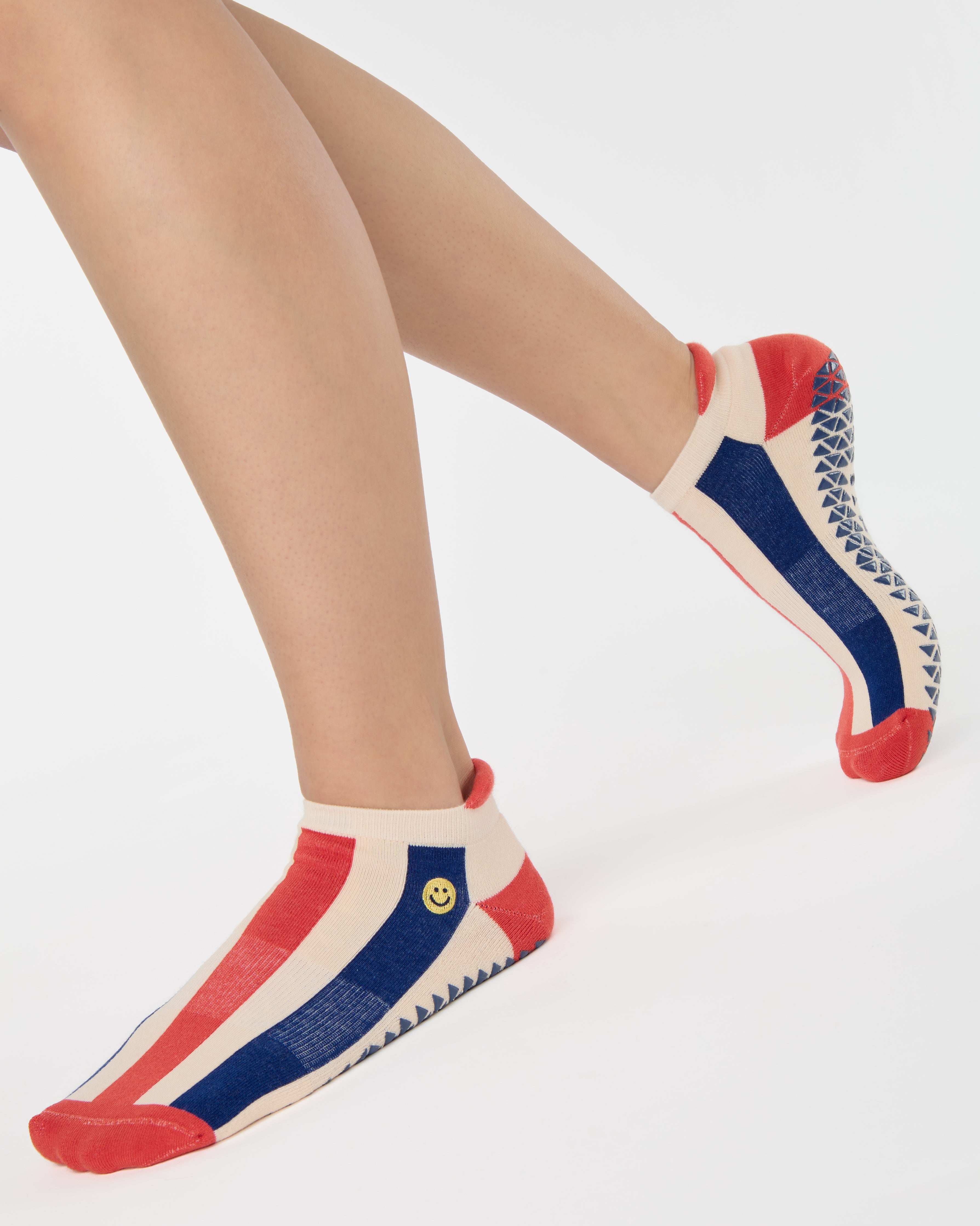 Woman wearing a playfully blue, white, and red stripped full foot grip sock with a little embroidered smiley face by the ankle. Made for pilates, barre, yoga, and beyond. 