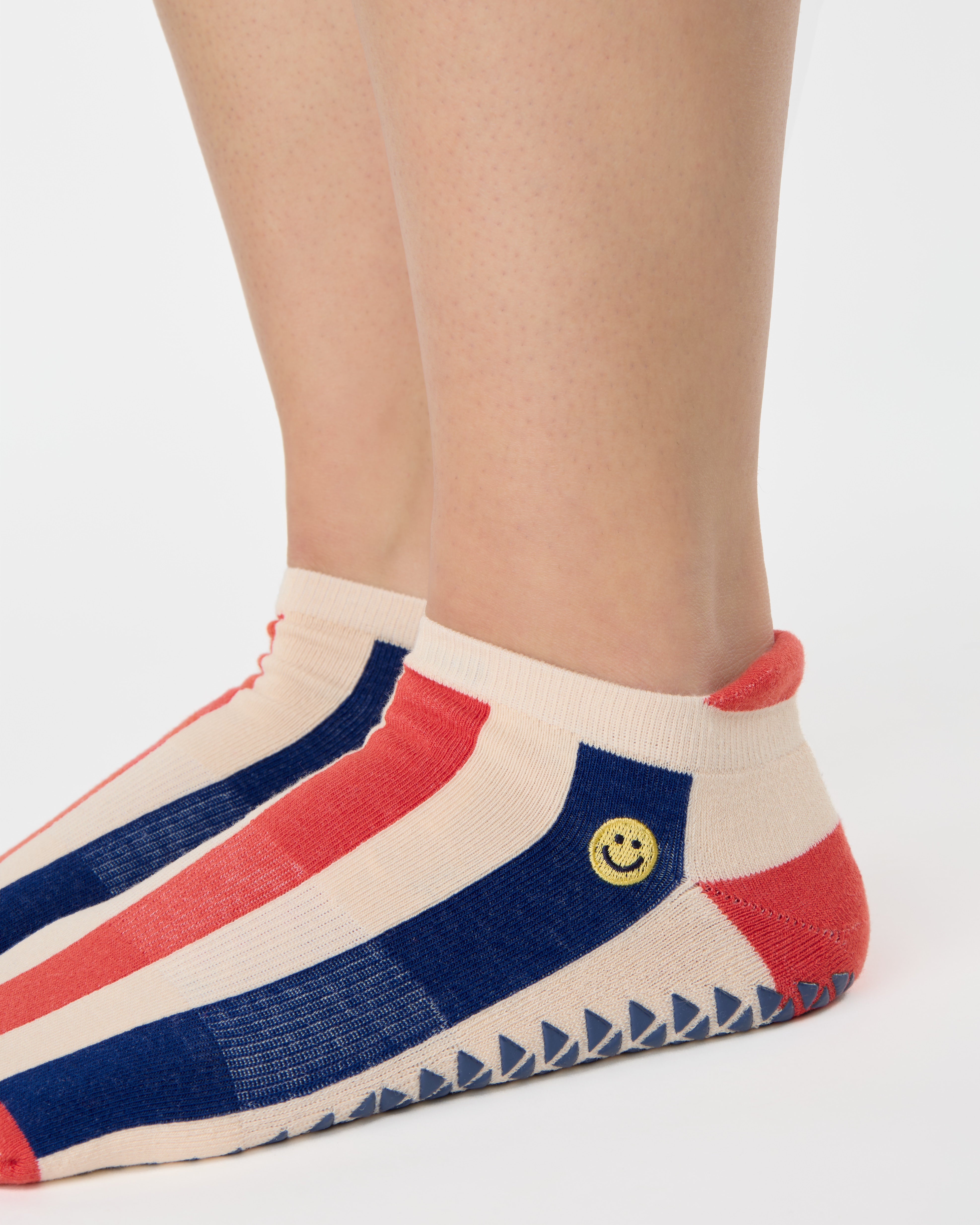 Woman wearing a playfully blue, white, and red stripped full foot grip sock with a little embroidered smiley face by the ankle. Made for pilates, barre, yoga, and beyond. 