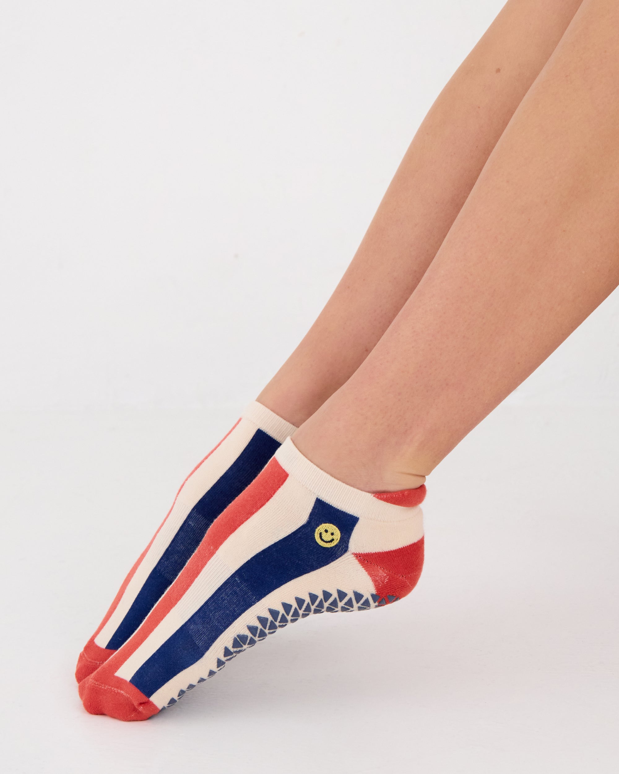Woman wearing a playfully blue, white, and red stripped full foot grip sock with a little embroidered smiley face by the ankle. Made for pilates, barre, yoga, and beyond. 
