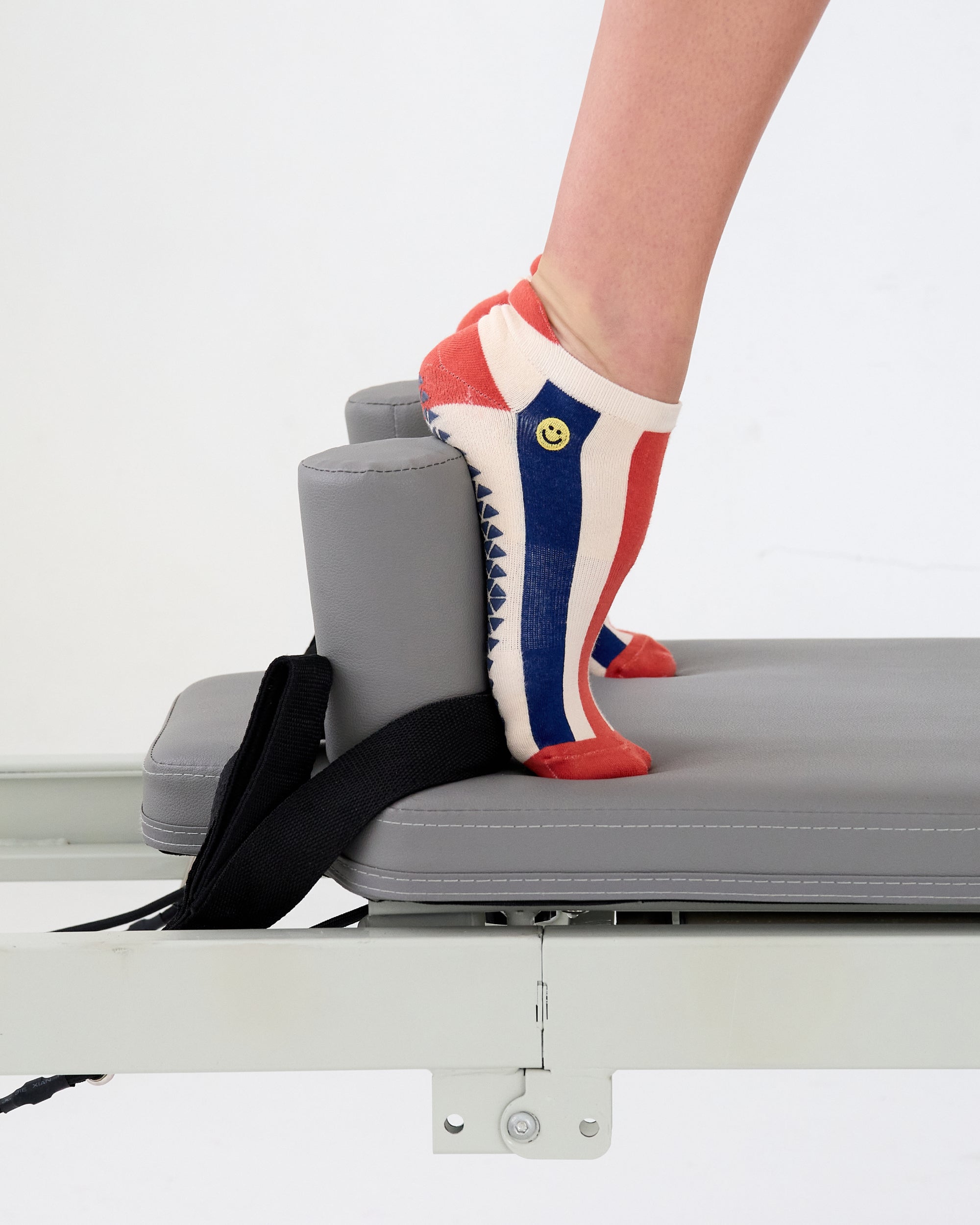 Woman on a pilates reformer wearing a playfully blue, white, and red stripped full foot grip sock with a little embroidered smiley face by the ankle. Made for pilates, barre, yoga, and beyond. 