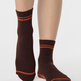 Woman wearing a solid chocolate brown ankle grip sock with orange stripes on the toes and top ankle cuff made for Pilates, Barre, Yoga, and beyond. 