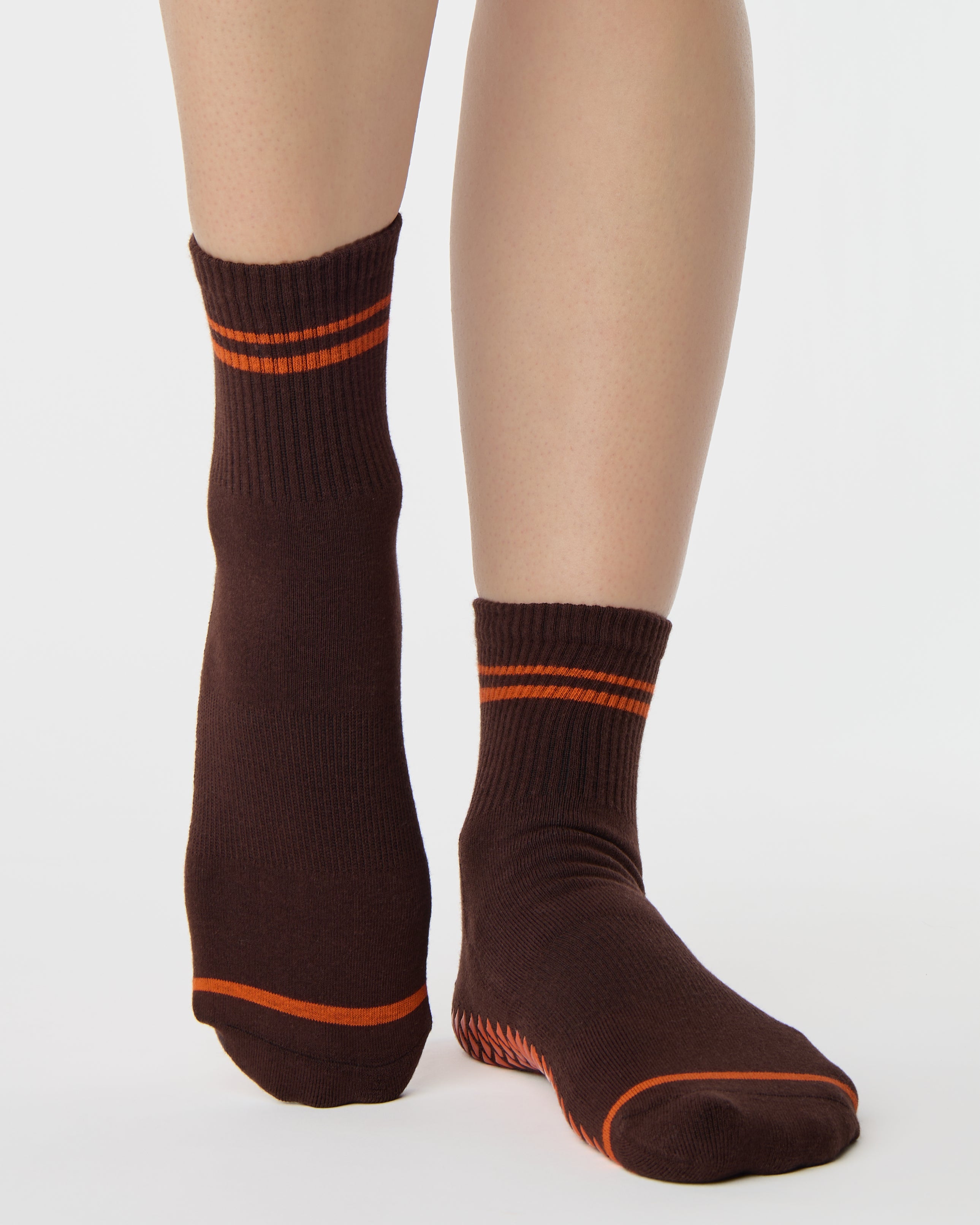 Woman wearing a solid chocolate brown ankle grip sock with orange stripes on the toes and top ankle cuff made for Pilates, Barre, Yoga, and beyond. 