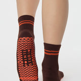 Woman wearing a solid chocolate brown ankle grip sock with orange stripes on the toes and top ankle cuff made for Pilates, Barre, Yoga, and beyond. 
