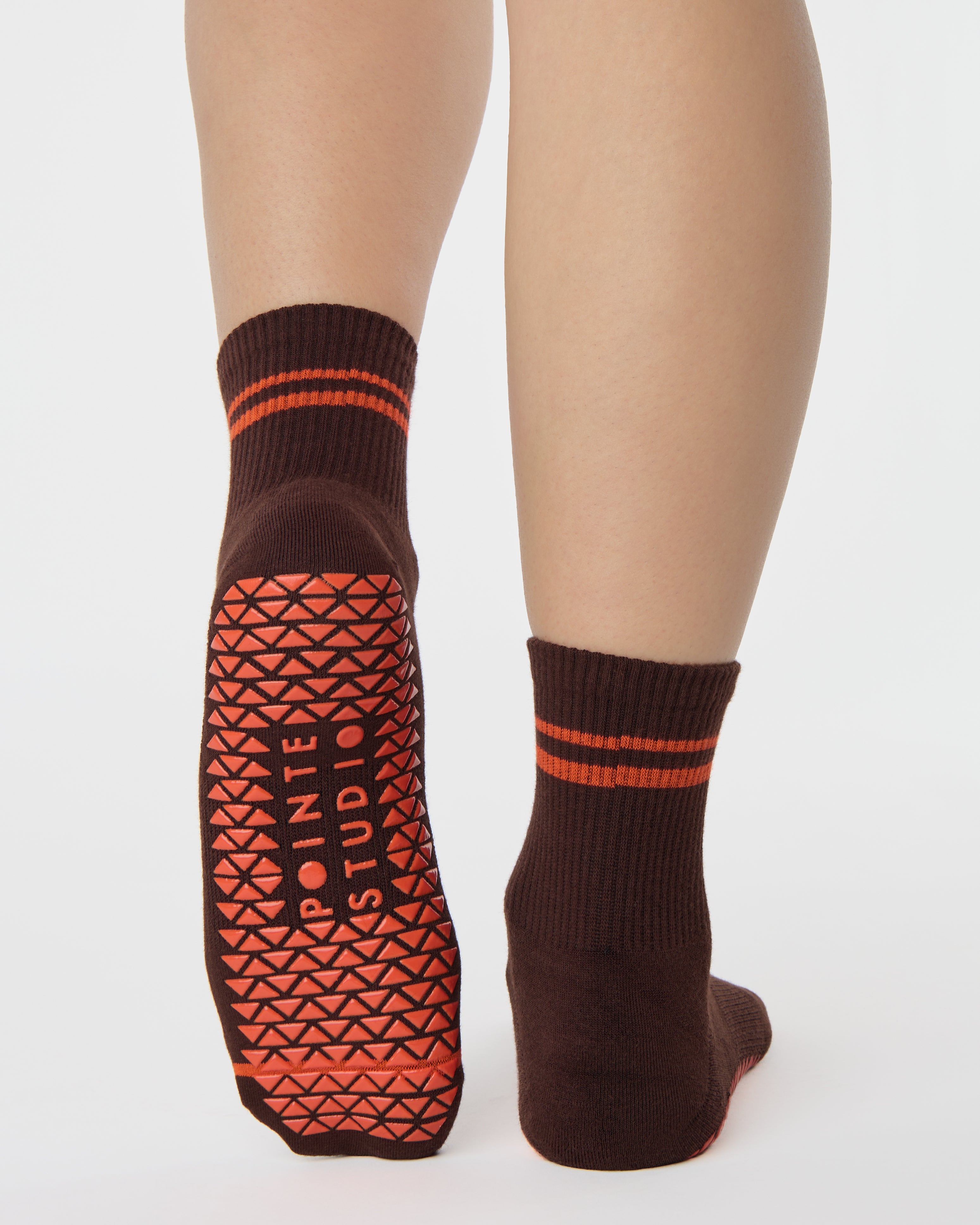 Woman wearing a solid chocolate brown ankle grip sock with orange stripes on the toes and top ankle cuff made for Pilates, Barre, Yoga, and beyond. 