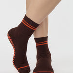 Woman wearing a solid chocolate brown ankle grip sock with orange stripes on the toes and top ankle cuff made for Pilates, Barre, Yoga, and beyond. 