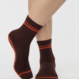 Woman wearing a solid chocolate brown ankle grip sock with orange stripes on the toes and top ankle cuff made for Pilates, Barre, Yoga, and beyond. 
