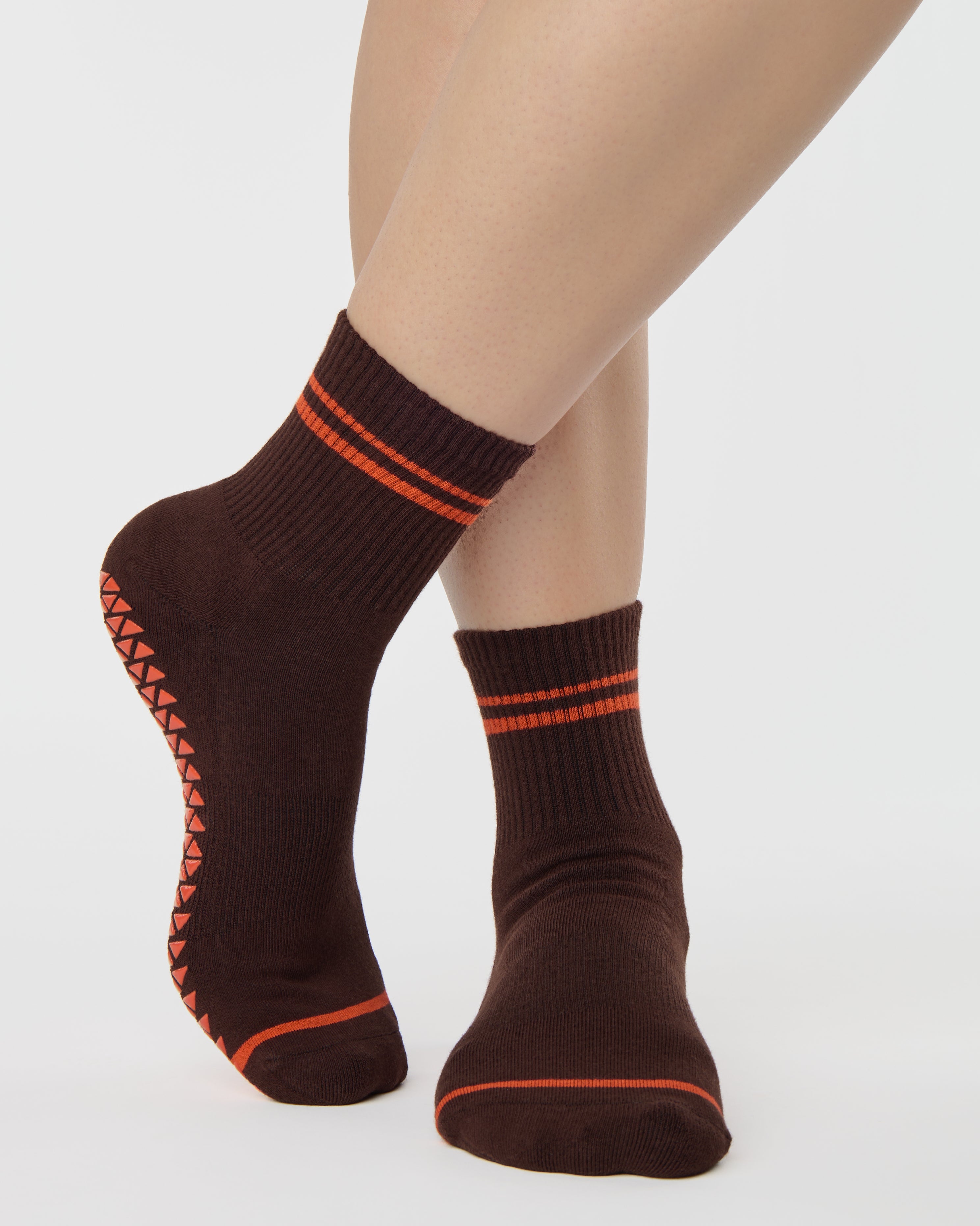 Woman wearing a solid chocolate brown ankle grip sock with orange stripes on the toes and top ankle cuff made for Pilates, Barre, Yoga, and beyond. 