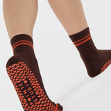 Woman wearing a solid chocolate brown ankle grip sock with orange stripes on the toes and top ankle cuff made for Pilates, Barre, Yoga, and beyond. 