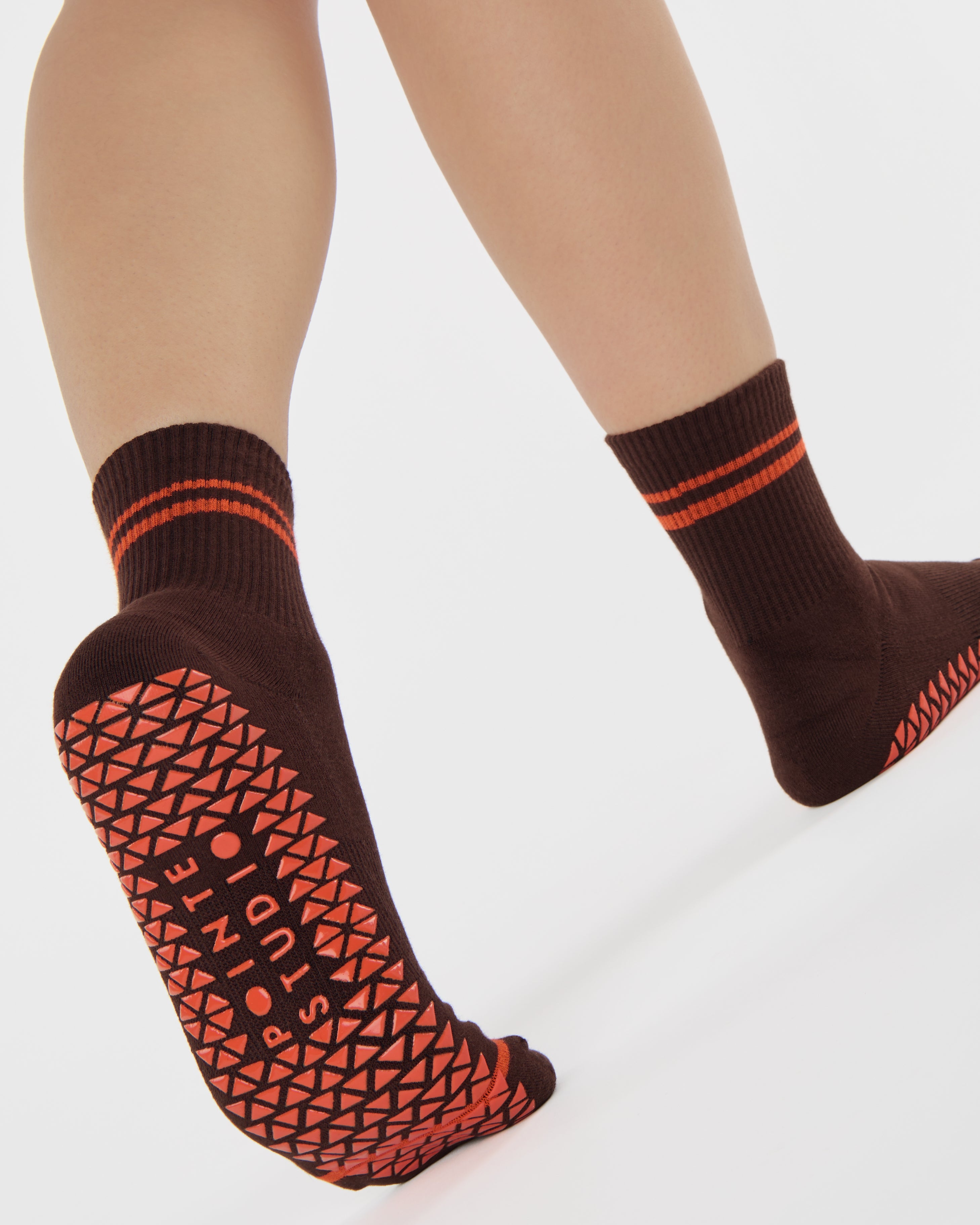 Woman wearing a solid chocolate brown ankle grip sock with orange stripes on the toes and top ankle cuff made for Pilates, Barre, Yoga, and beyond. 