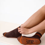 Woman on yoga mat wearing a solid chocolate brown ankle grip sock with orange stripes on the toes and top ankle cuff made for Pilates, Barre, Yoga, and beyond. 