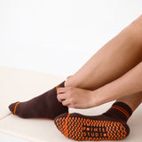 Woman on yoga mat wearing a solid chocolate brown ankle grip sock with orange stripes on the toes and top ankle cuff made for Pilates, Barre, Yoga, and beyond. 