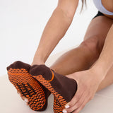 Woman on yoga mat wearing a solid chocolate brown ankle grip sock with orange stripes on the toes and top ankle cuff made for Pilates, Barre, Yoga, and beyond. 