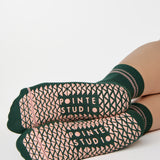 Woman wearing a solid green ankle grip sock with pink stripes on the toes and top ankle cuff made for Pilates, Barre, Yoga, and beyond. 