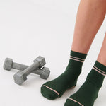 Woman wearing a solid green ankle grip sock with pink stripes on the toes and top ankle cuff made for Pilates, Barre, Yoga, and beyond. 